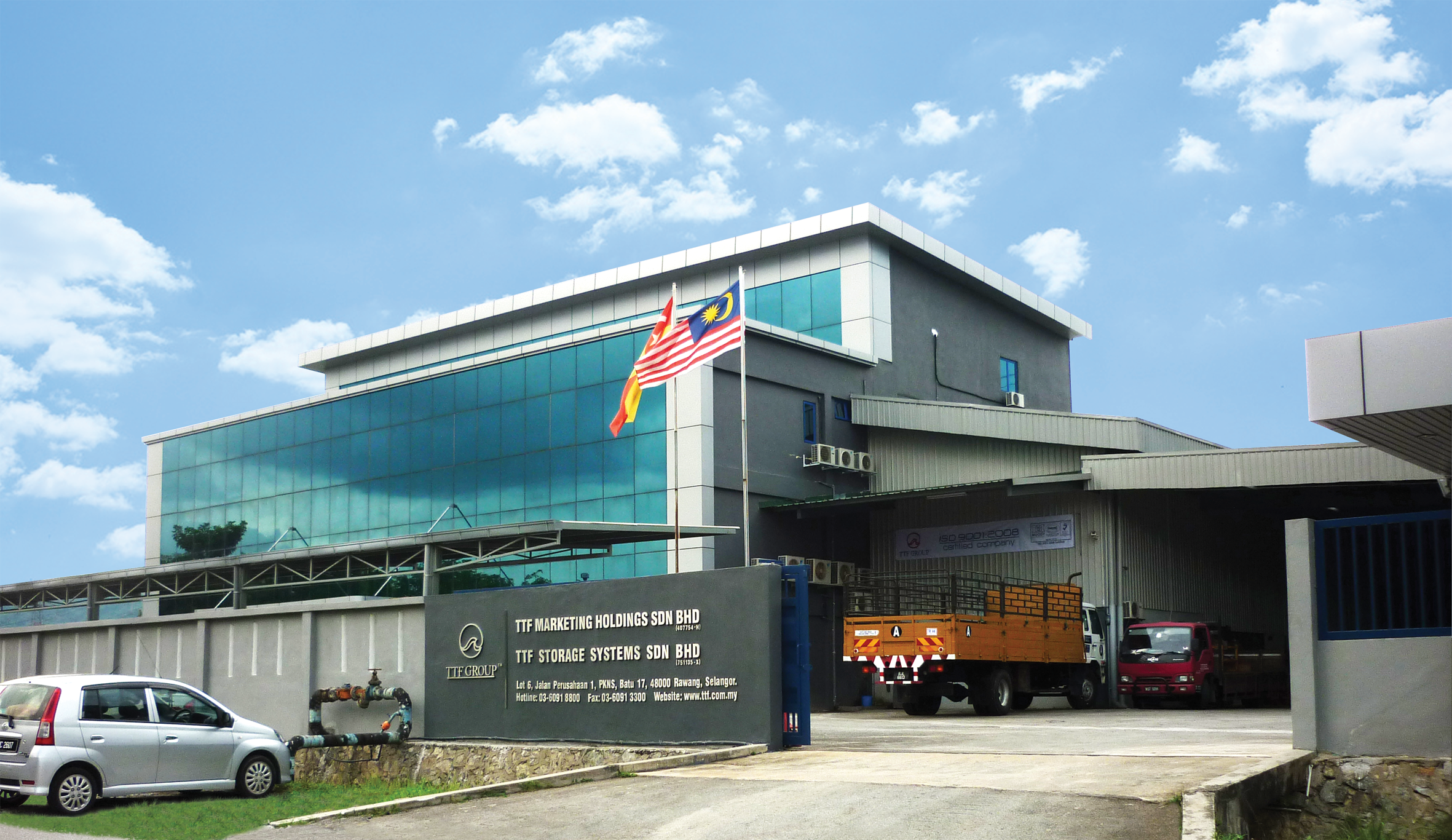 TTF Marketing Holdings Sdn Bhd and TTF Storage Systems Sdn Bhd office building at TTF Group - Rawang HQ, Kawasan Perusahaan PKNS, Batu 17, Rawang, Selangor, Malaysia. The modern industrial building features blue glass facade and grey warehouse sections. The Malaysian flag is visible in front.