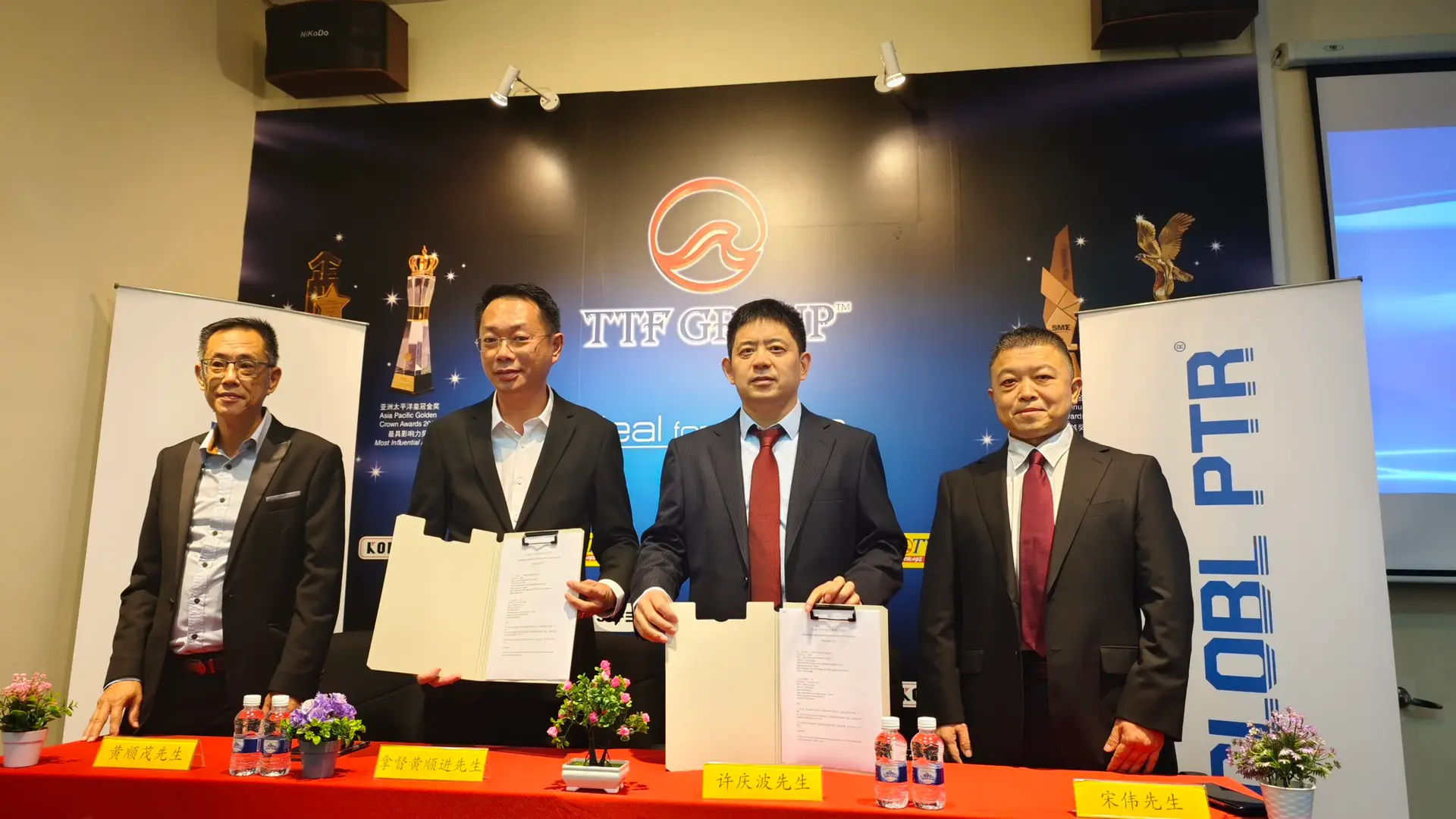 TTF Group signing ceremony: Four formally dressed Asian businessmen holding signed documents and smiling for a photo. The TTF Group logo and awards are visible in the background, highlighting a successful business agreement.